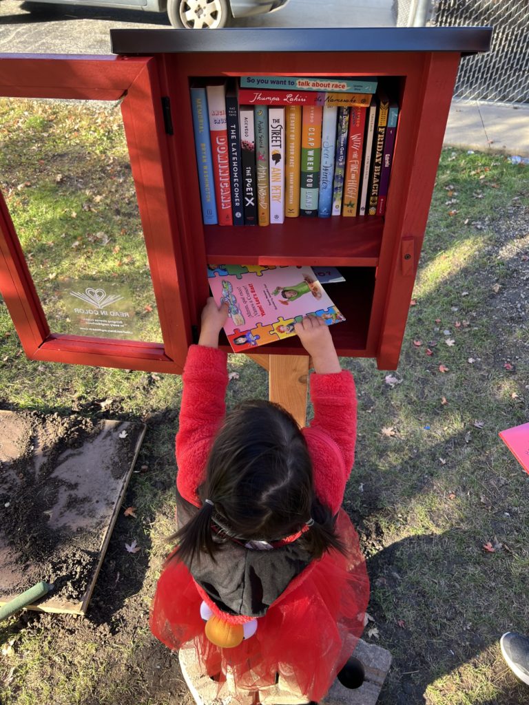 Small dark haired girl reaches up to put a book in a red framed bookbox on a red post.